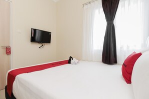 Ein Hotelzimmer mit Bett, Wandfernseher, Fenster mit Vorhängen und roter Bettdecke.