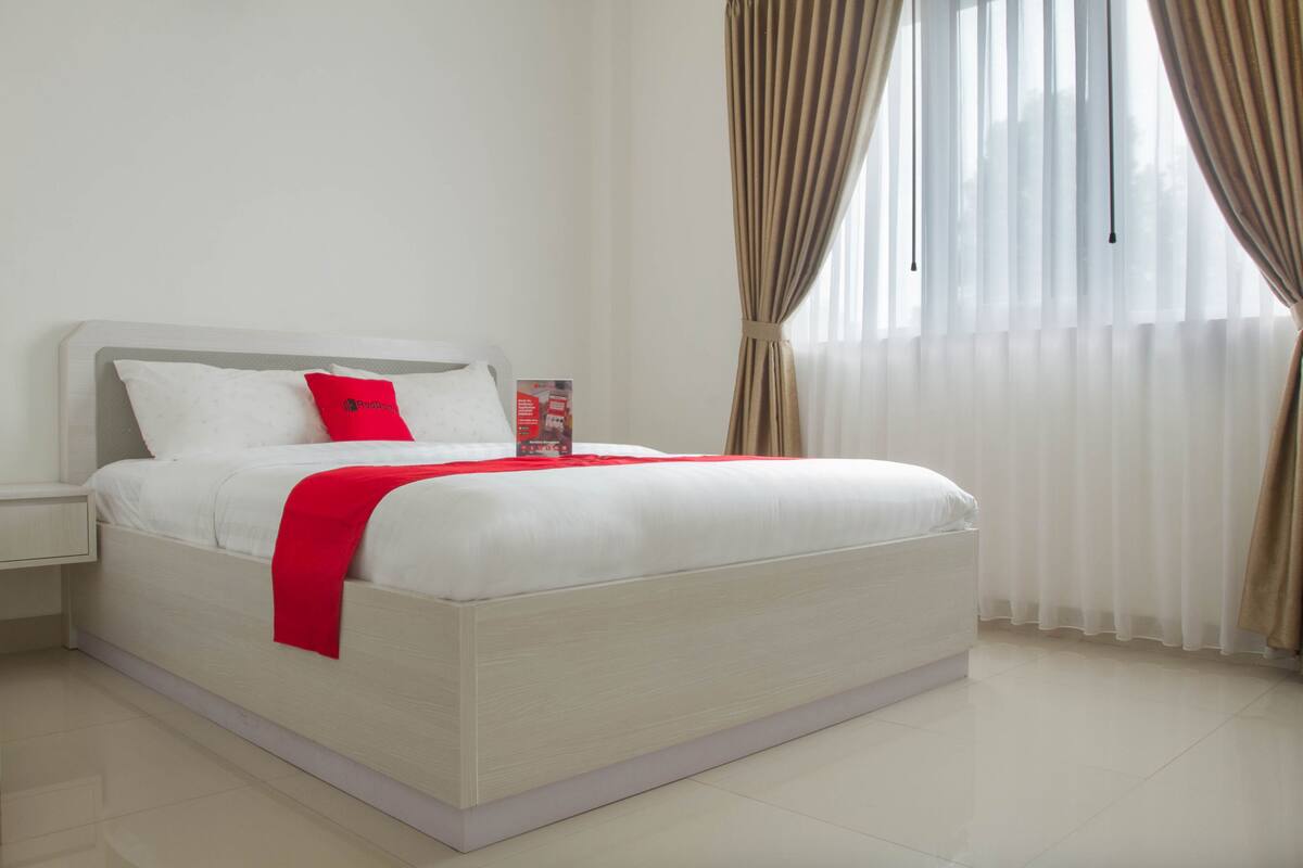 A neatly made bed with white bedding and a red accent, a bedside table, and a window with sheer curtains.