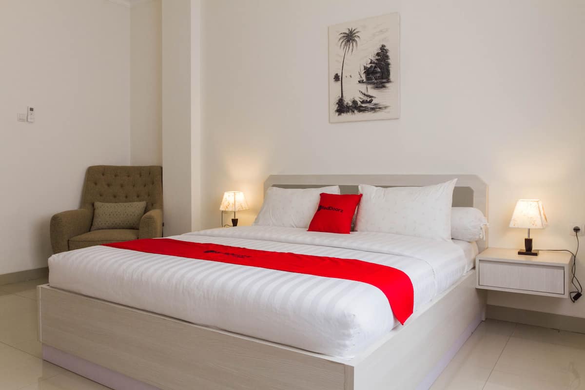 A neatly made bed with a red accent pillow, a chair, and a nightstand with a lamp.