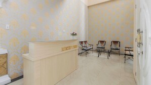 A reception area with a wooden reception desk, patterned wallpaper, and four chairs arranged in a row. - RedDoorz near Grand Palladium (Medan)
