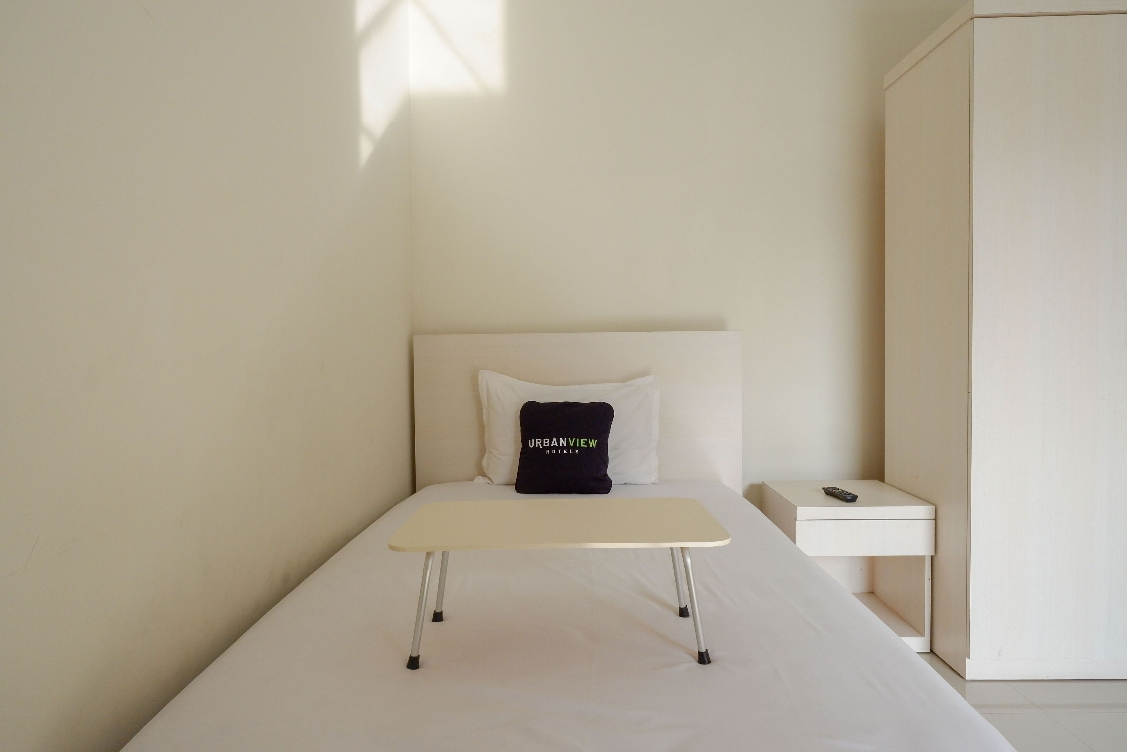 Ein schlicht eingerichtetes Hotelzimmer mit einem Einzelbett, einem kleinen Nachttisch und einem Kopfkissen mit dem Namen des Hotels.