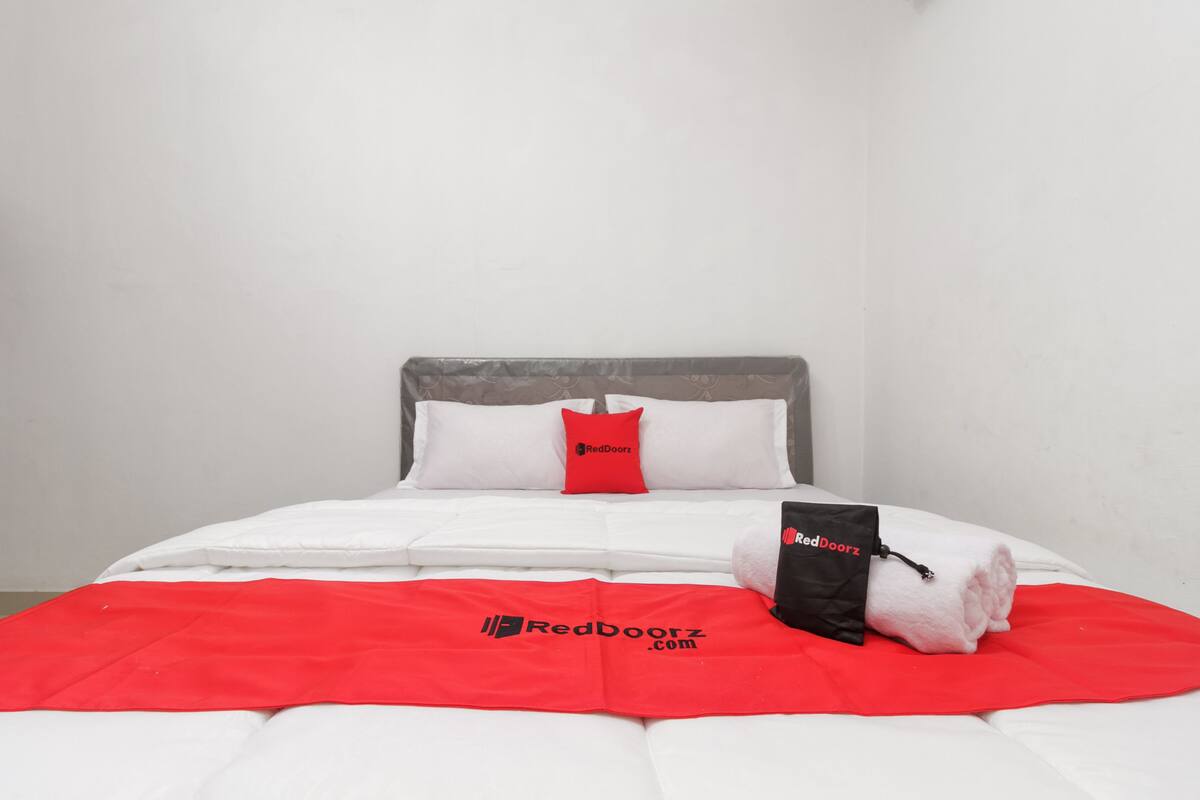 A neatly made bed with a red blanket and pillows, a rolled towel, and a RedDoorz branded bag.