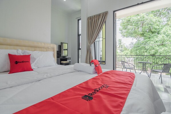 Une chambre d’hôtel comprenant un lit, une couverture de la marque RedDoorz, un oreiller rouge, une table de chevet, une fenêtre avec des rideaux et un balcon avec des meubles d’extérieur.