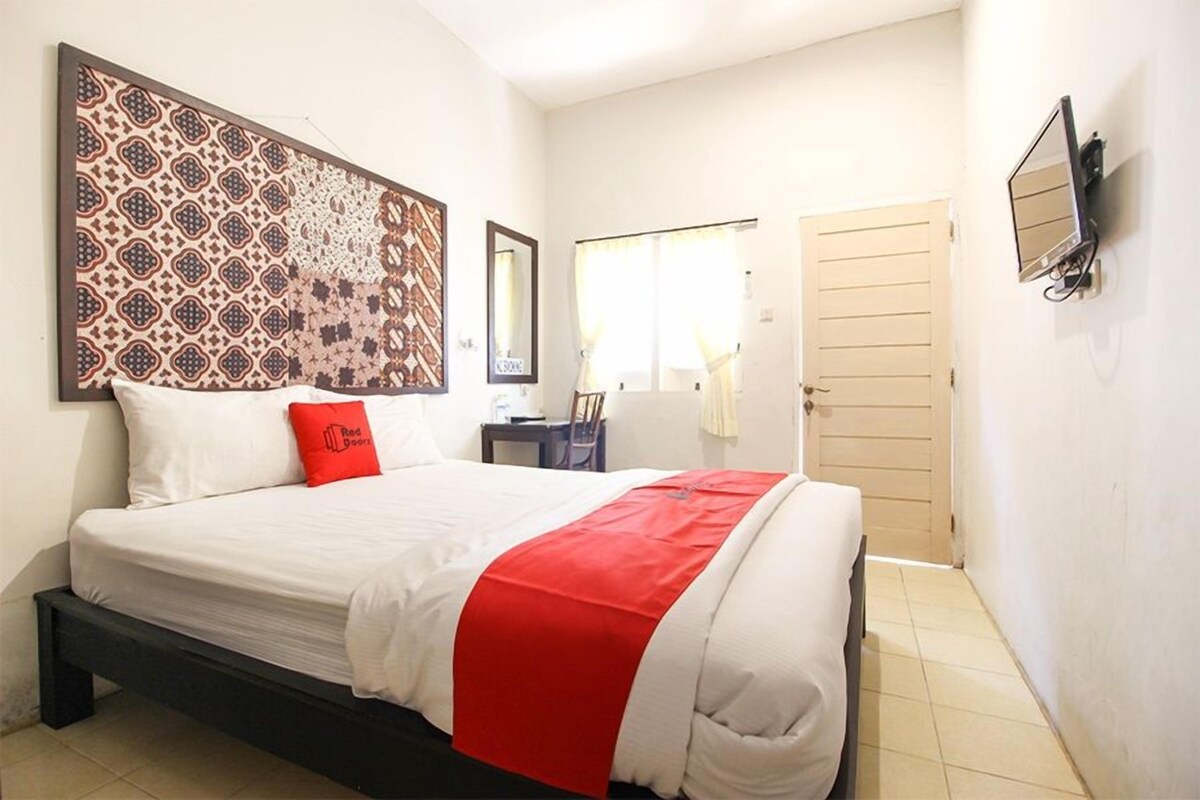Une chambre d’hôtel avec un lit, un plaid rouge, une grande décoration murale à motifs, un miroir, une télévision et une porte.