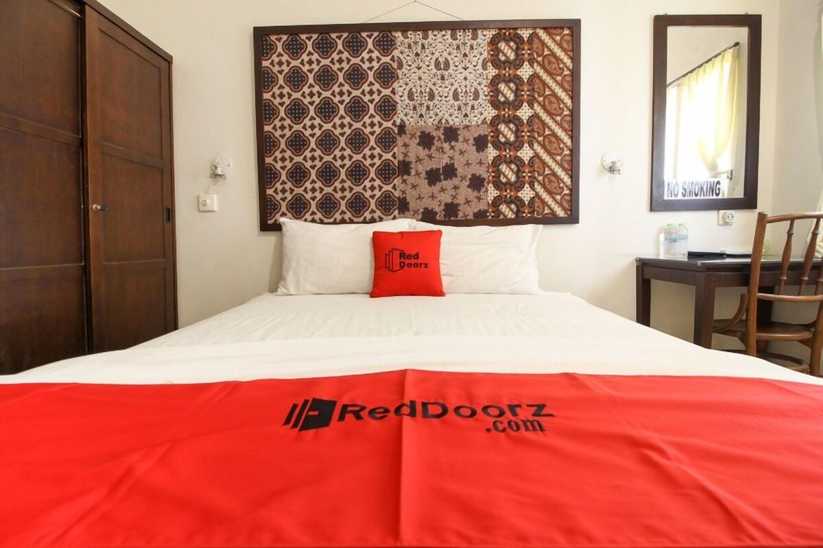 Une chambre d’hôtel avec un lit, un oreiller rouge, une tête de lit à motifs, une armoire en bois, une chaise et un panneau « Il est interdit de fumer ».