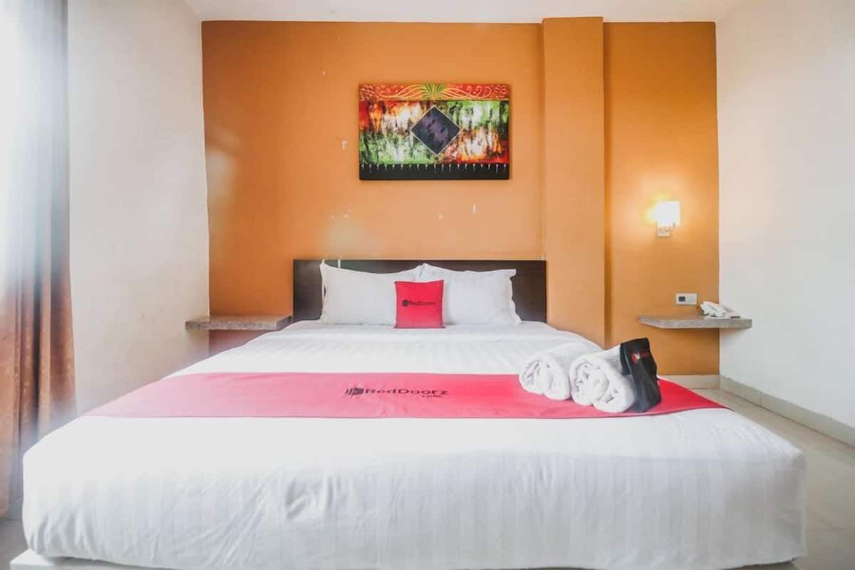 A hotel room with a bed, a nightstand, a wall-mounted artwork, a red bedspread, and a towel on the bed.