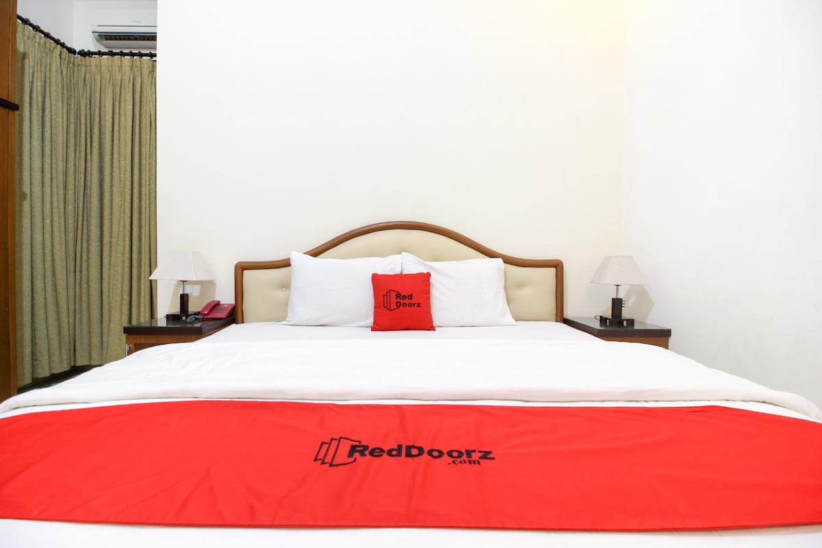 A hotel room with a bed, a nightstand with a lamp, a telephone, and a red pillow with the RedDoorz logo.