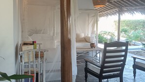 A room with a thatched roof, a bed with white linens, a wooden chair, and a small table with books. - Pole Pole Beach House (Watamu)