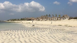 A sandy beach with footprints, a clear sky, and a row of thatched-roof buildings in the background. - Pole Pole Beach House (Watamu)