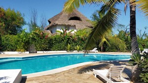 A swimming pool with a thatched-roof building in the background, surrounded by lush greenery and palm trees. - Pole Pole Beach House (Watamu)