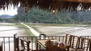 Een eethoek met bamboemeubels, een tafel gedekt met borden, kommen en flessen, met uitzicht op een rivier en groen.
