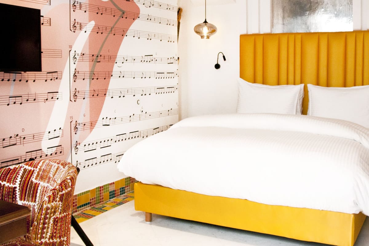 A bedroom with a yellow bed, white bedding, a patterned chair, and a wall with musical notes and the word "satisfaction".