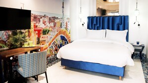 A hotel room with a bed, a desk, a chair, a TV, and a colorful mural on the wall.