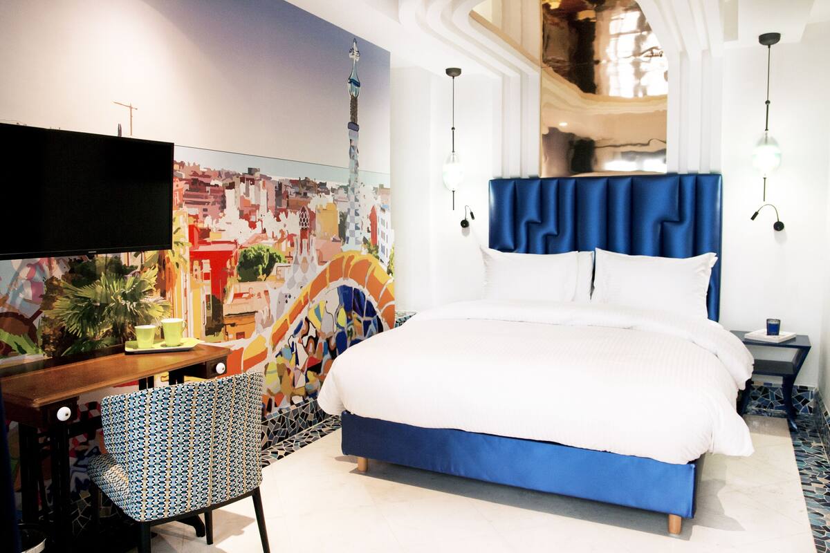 A hotel room with a bed, a desk, a chair, a TV, and a colorful mural on the wall.