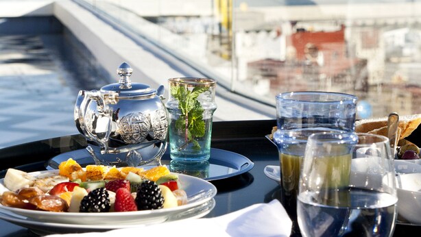 A table with a fruit platter, a teapot, and glasses of water and juice, with a view of a marina and boats in the background.