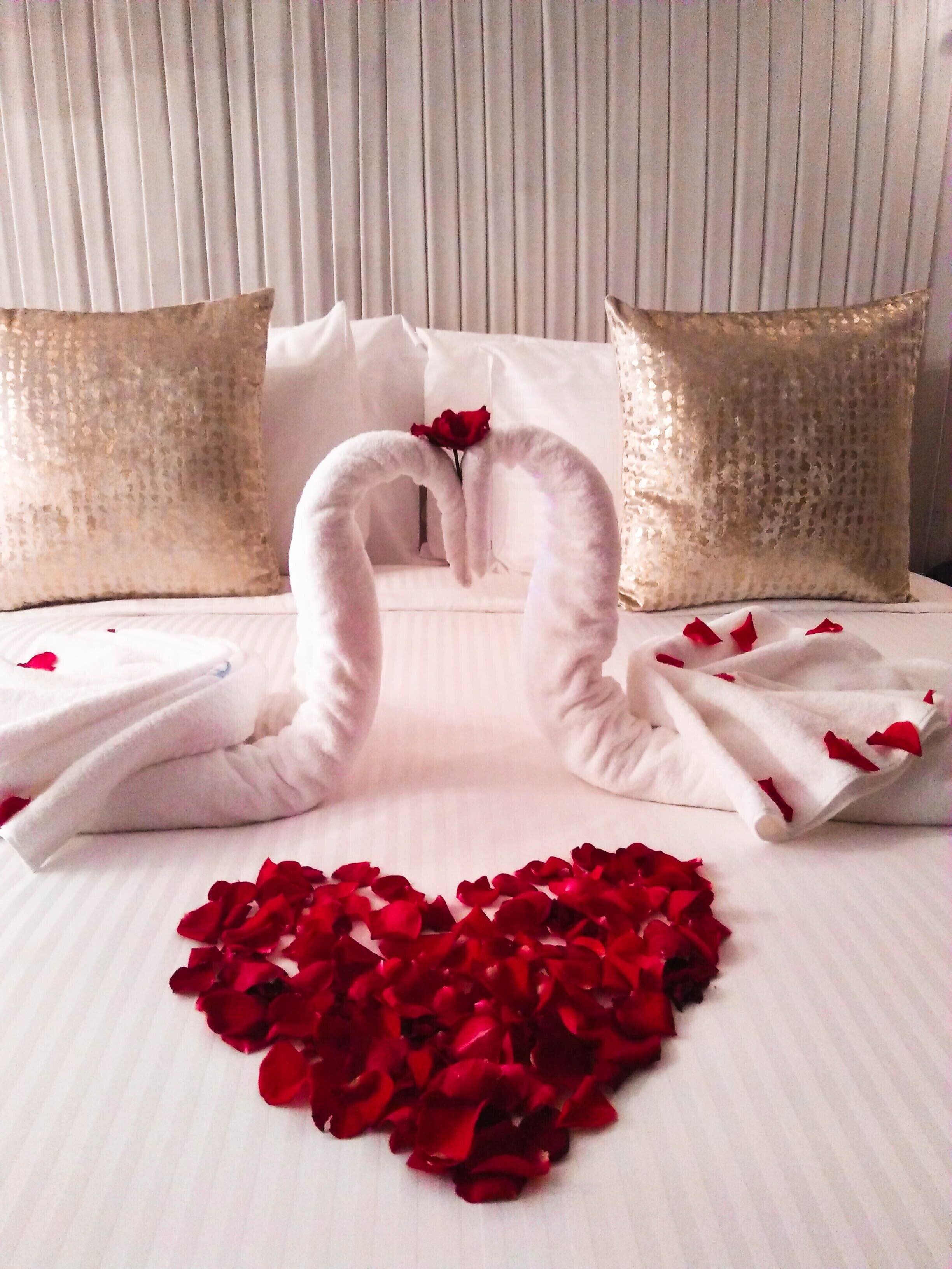 A bed with a heart-shaped arrangement of rose petals and two swan-shaped towels.
