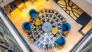 A spacious atrium with a geometric patterned floor, a central seating area with blue and yellow chairs, and large windows with warm lighting.