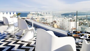 A rooftop dining area with modern white chairs and a glass table, overlooking a cityscape and the sea.
