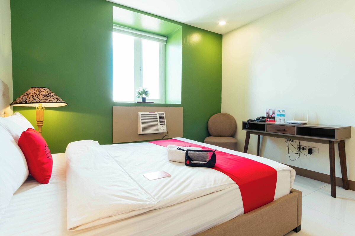 A hotel room with a bed, a desk, a chair, a lamp, and a window.