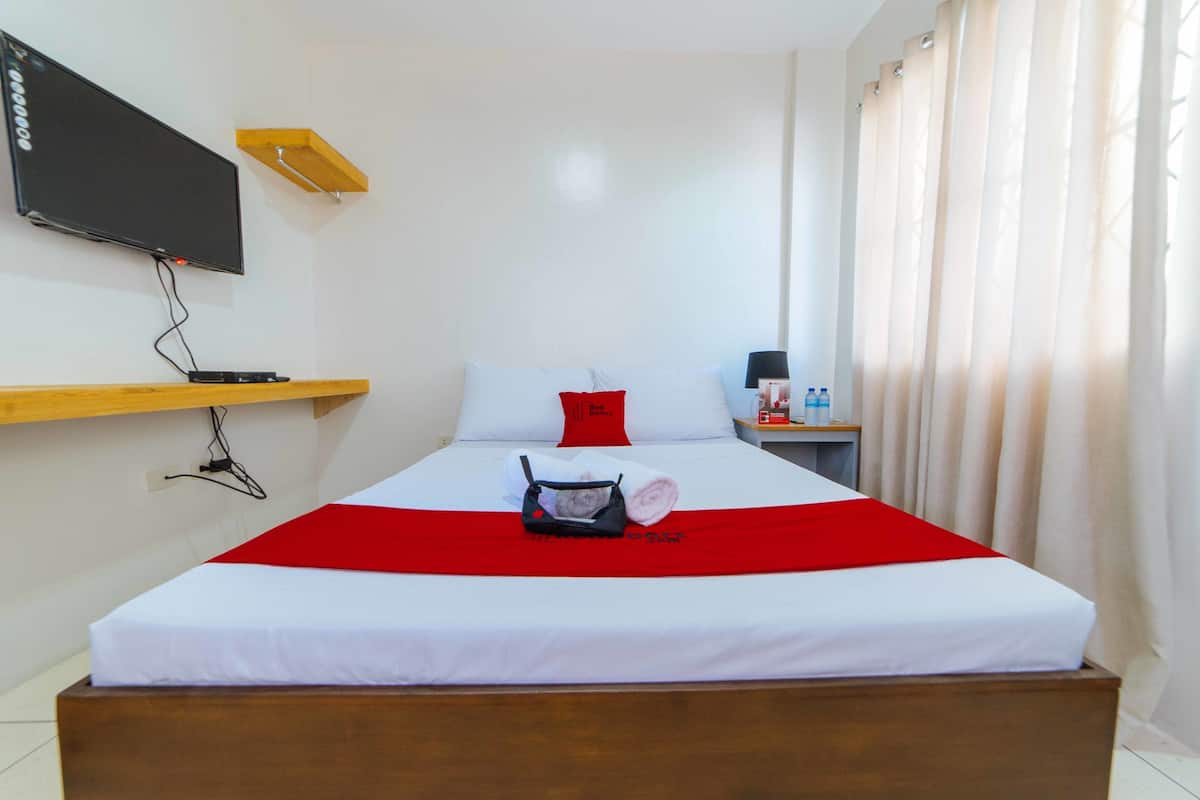 Une chambre d’hôtel avec un lit, un téléviseur fixé au mur, une étagère en bois, une table de chevet avec une lampe et une fenêtre avec des rideaux.