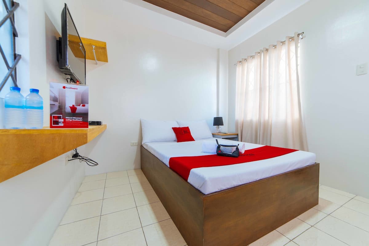 Une chambre d’hôtel avec un lit en bois, du linge de lit blanc, un plaid rouge, une table de chevet avec une lampe et un téléviseur fixé au mur.
