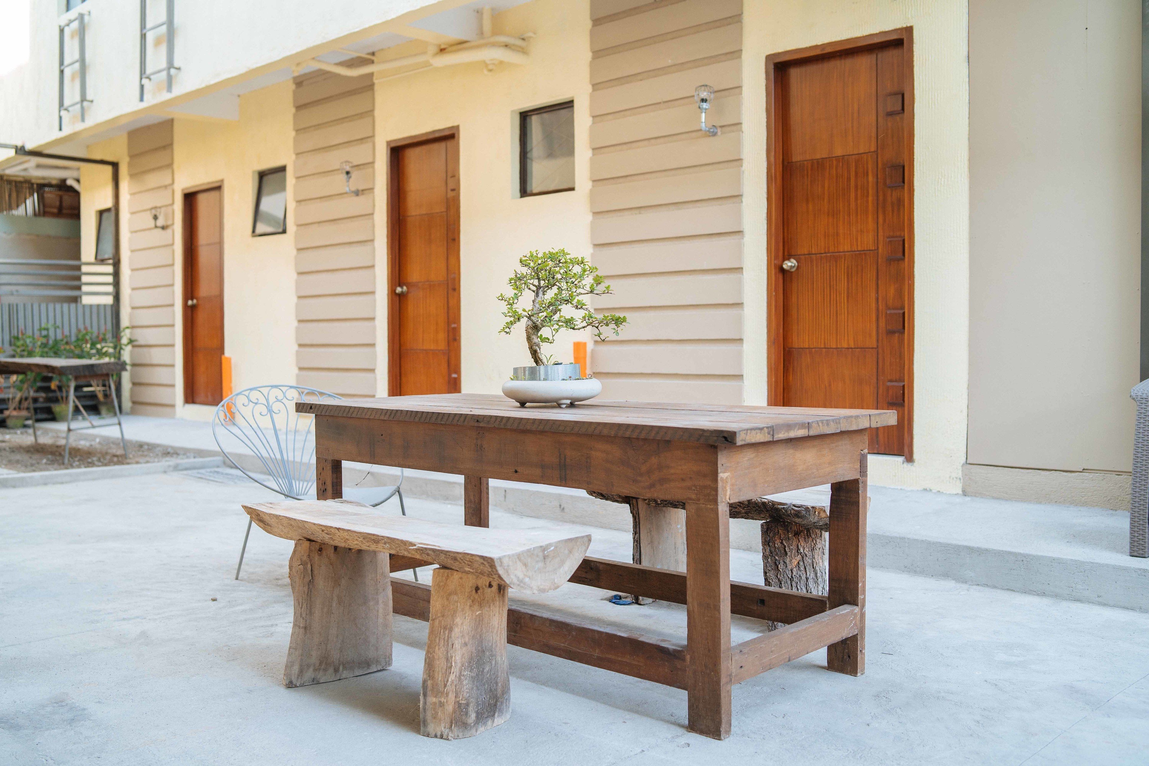 An outdoor patio with a wooden table and bench, a potted plant, and a building with beige siding and wooden doors.