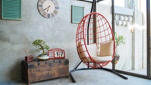 Une pièce comprenant un coffre en bois, une chaise en fil de fer rouge, une horloge murale et une plante en pot.