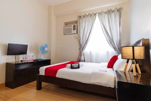 Une chambre d’hôtel avec un lit, une télévision, une table de chevet avec une lampe, un ventilateur et une fenêtre avec des rideaux.