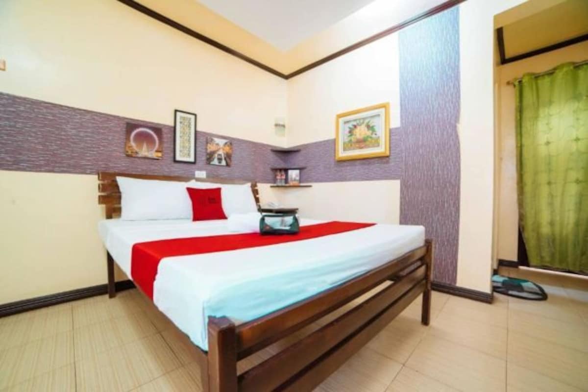 A neatly made bed with a red and white bedspread, a wooden headboard, and a small bedside table with a lamp. The room has a tiled floor and a green curtain on the window.