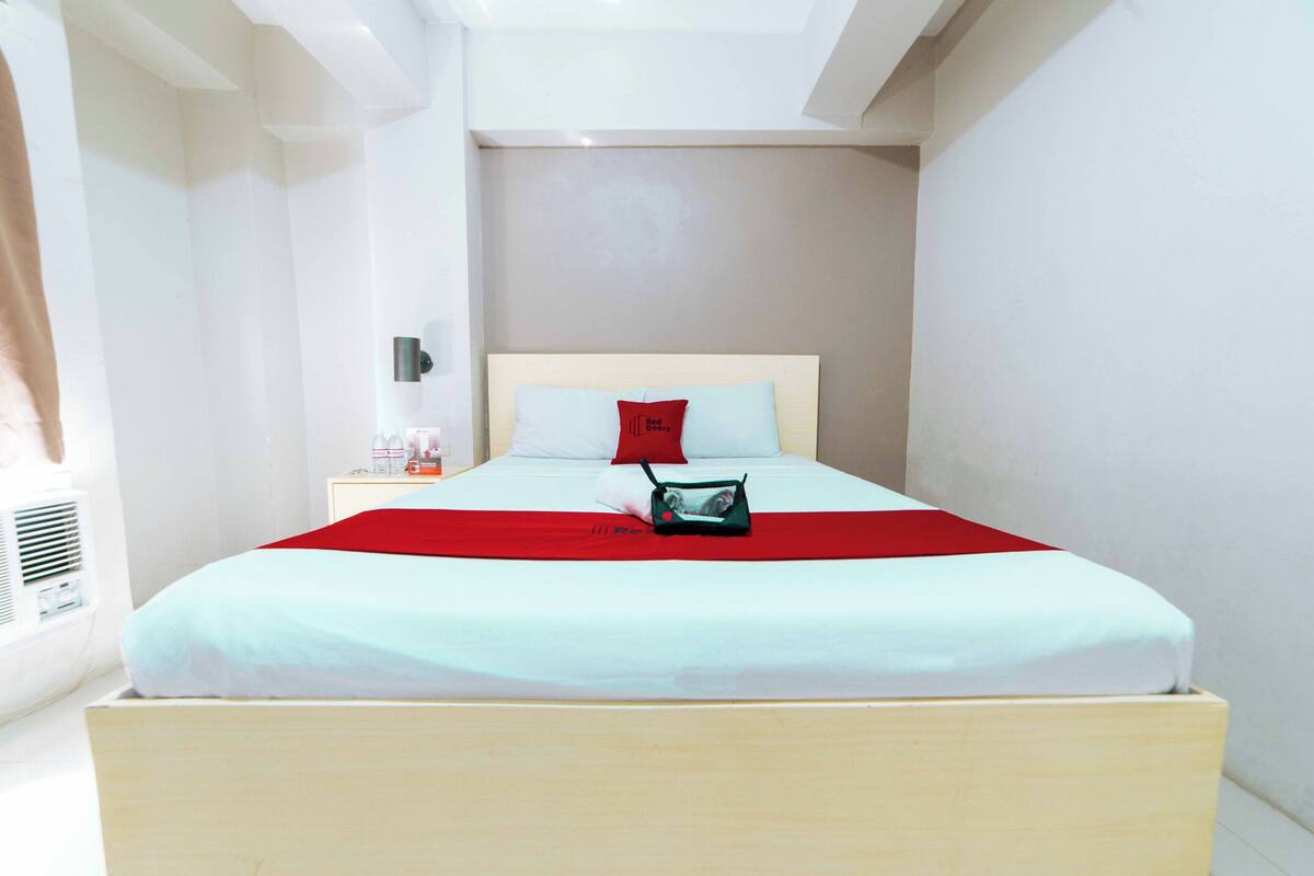 A neatly made bed with a red blanket, a small bedside table with a lamp, and a wall-mounted air conditioner.