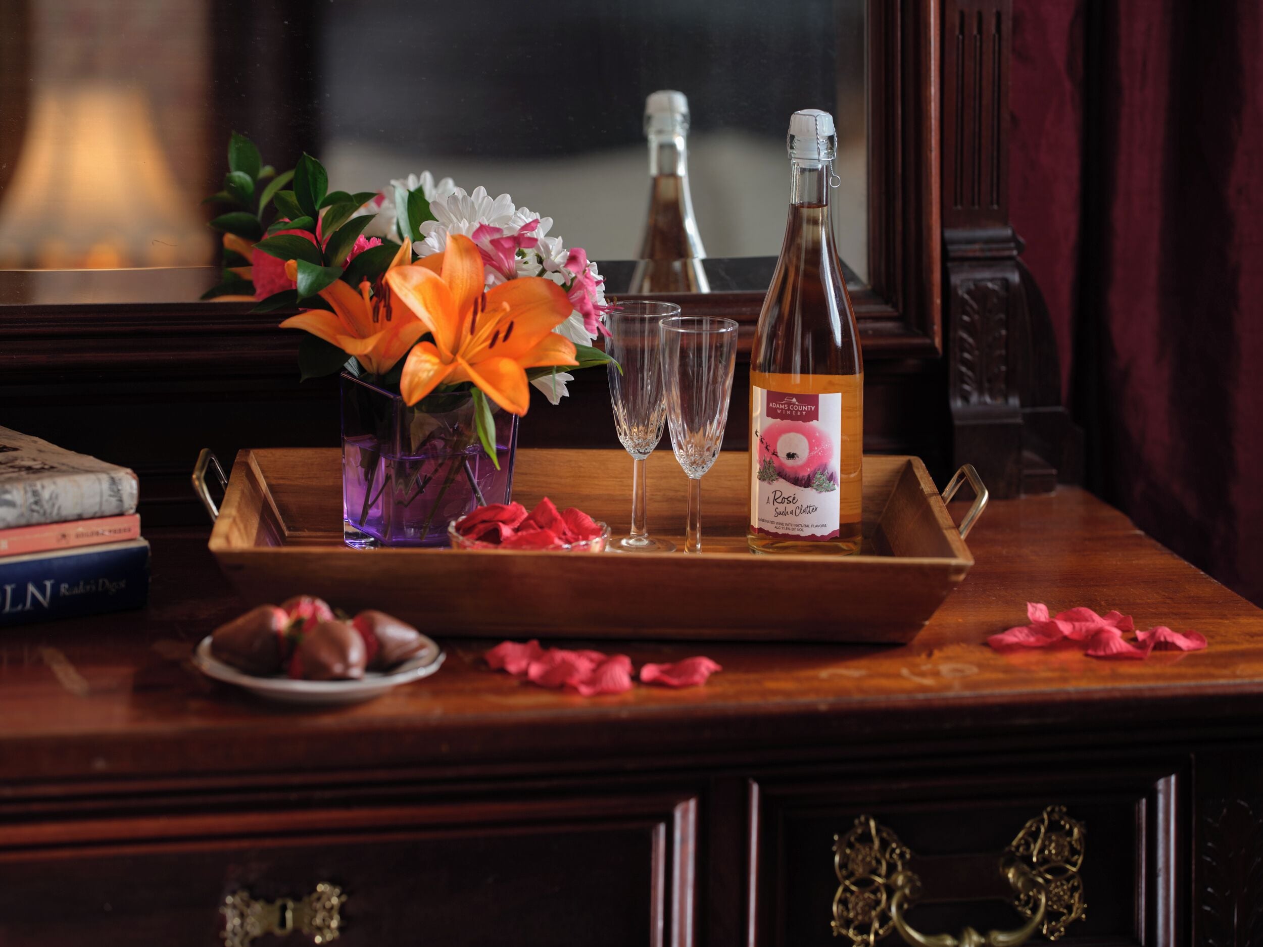 A wooden tray with a bottle of rose syrup, two glasses, a vase with flowers, and scattered rose petals on a wooden table.