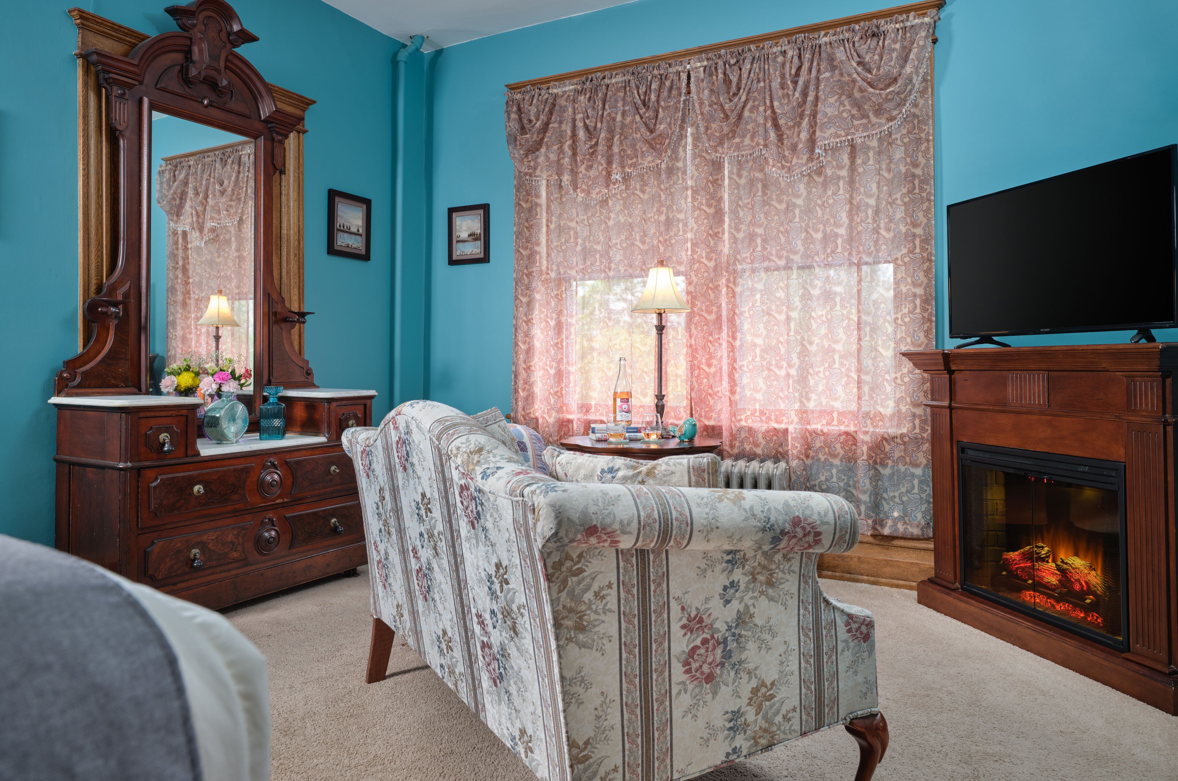 Une pièce avec un mur bleu, une commode en bois, un canapé à motifs floraux, un miroir, une cheminée et un téléviseur.