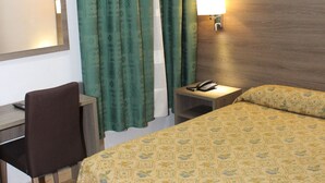 A hotel room with a bed, a desk, a chair, a nightstand with a lamp, and a window with curtains. - Hotel Nacional (Melilla)
