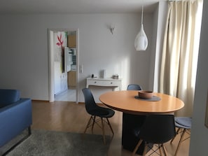 Ein modernes Wohnzimmer mit einem blauen Sofa, einem runden Holzesstisch mit vier Stühlen, einem weißen Schreibtisch und einer hängenden Pendelleuchte.