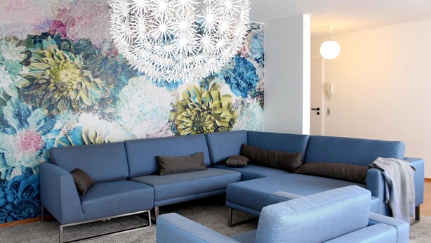 Een moderne woonkamer met een blauwe modulaire bank, een decoratief wandtapijt en een hanglamp.