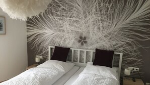 Een bed met wit beddengoed en donkere kussens, een wit hoofdbord, en een muur met een decoratief veerdessin.