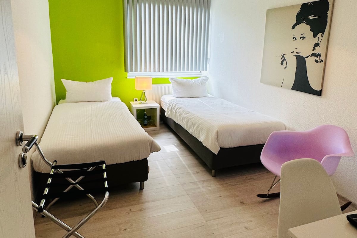 Una habitación de hotel moderna con dos camas, un muro verde decorativo, un ventanal con persianas, una lámpara en el techo y un cuadro en la pared.