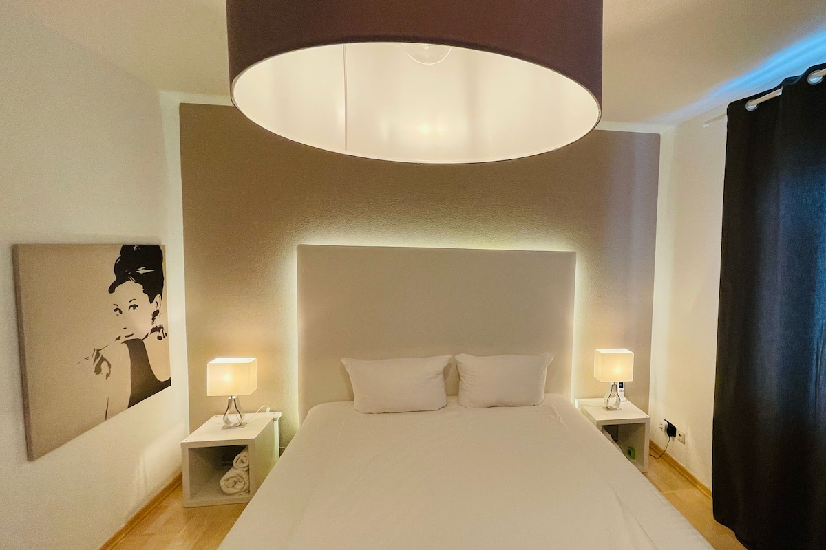 Habitación de hotel con una cama grande, dos mesitas de noche con lámparas, una obra de arte colgada en la pared y una cortina oscura.