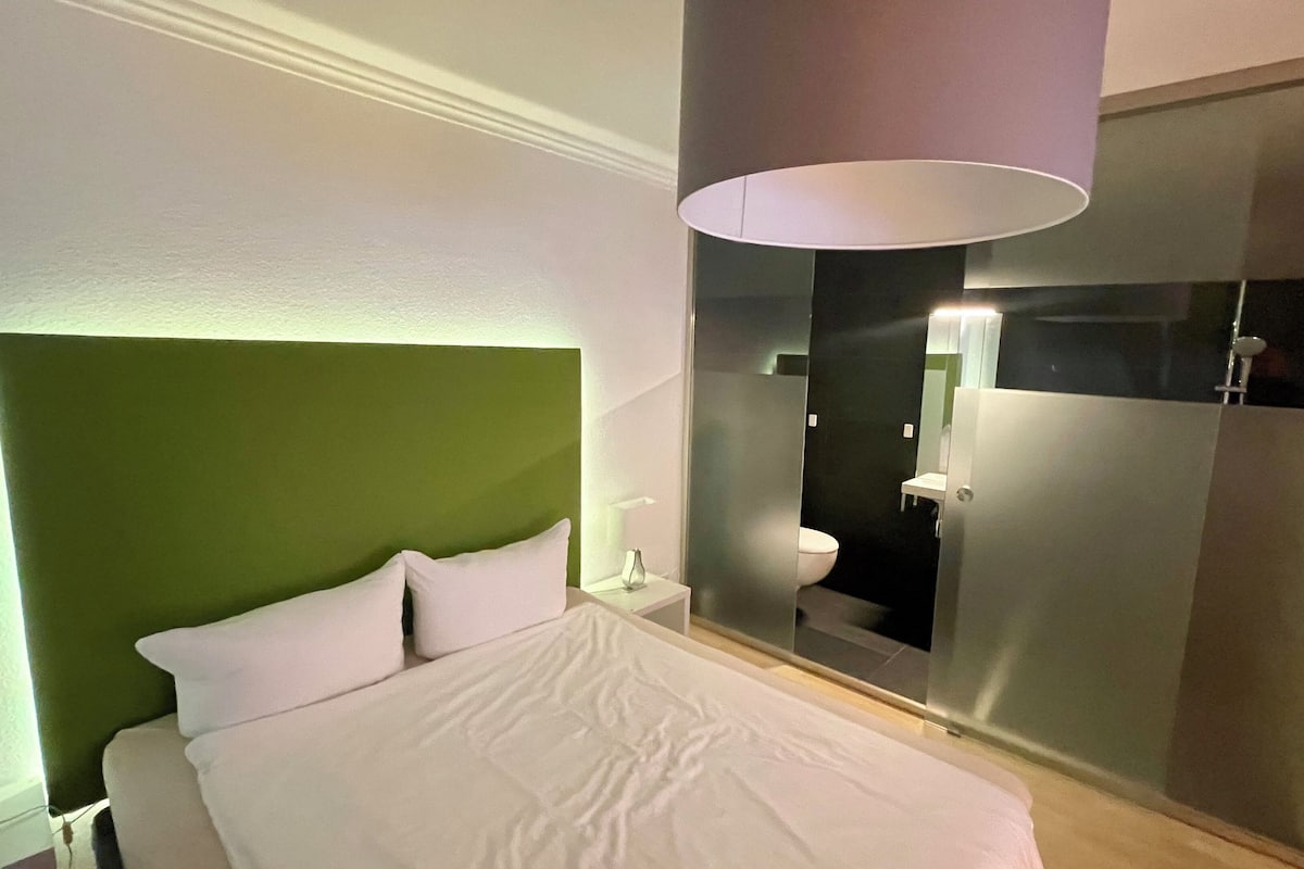 Una habitación de hotel moderna con una cama grande, una mesita de noche, una lámpara de noche, una pared de acento verde, un separador de vidrio y un baño con inodoro y lavabo.