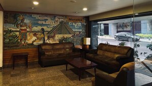A hotel lobby with a mural depicting ancient figures and a pyramid, leather sofas, a coffee table, and a glass wall offering a view of the street. - Hotel Manalba (Mexico City)