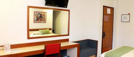Habitación de hotel con escritorio de madera, una silla roja, cama con ropa de cama verde, televisor montado en la pared y una puerta con un letrero.