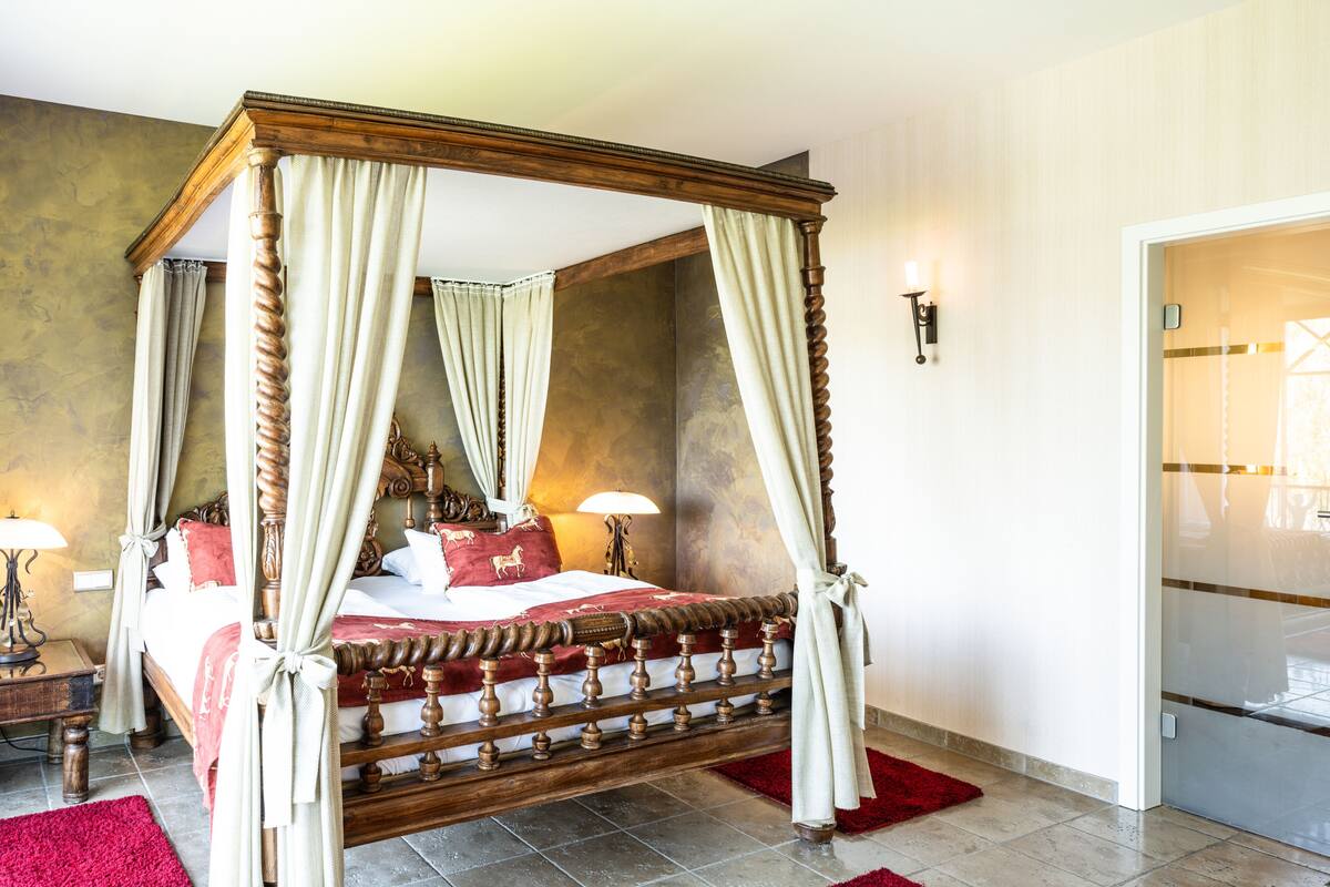 A four-poster bed with a canopy, a nightstand with a lamp, and a red rug on a tiled floor.