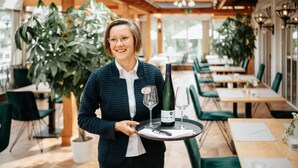 A person holding a tray with a bottle and two glasses, in a restaurant with wooden tables and green chairs, and large potted plants. - Wellness- & Genusshotel Weisser Bär (Muelheim)