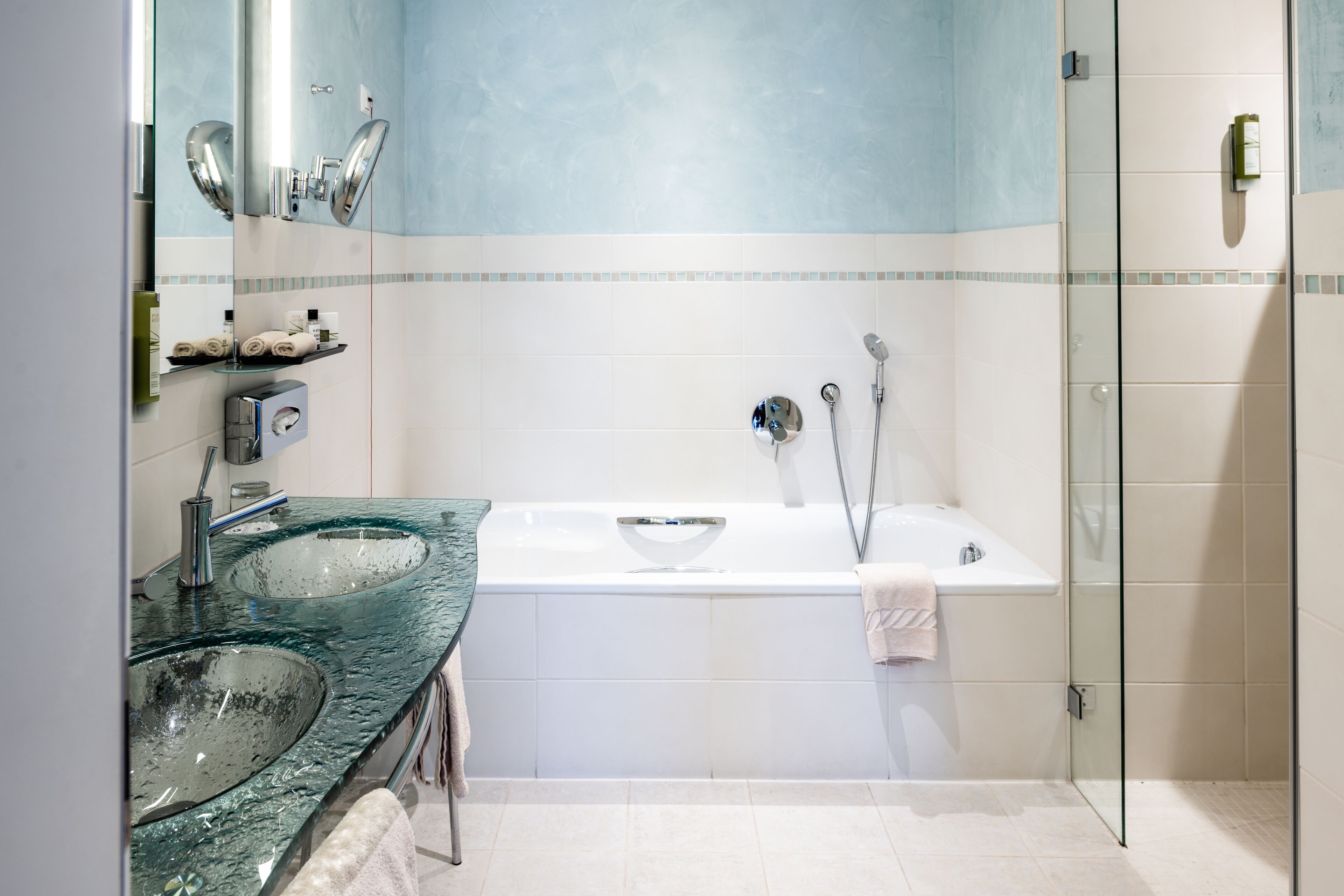 A modern bathroom with two glass sinks, a white bathtub, and a glass-enclosed shower.