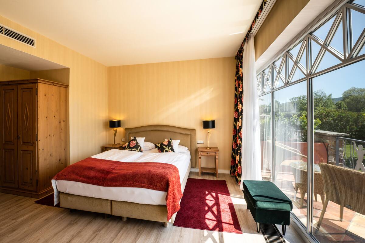 A bedroom with a large bed, a wooden wardrobe, a nightstand with a lamp, and a view of the outdoors through large windows.