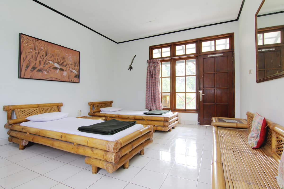 A room with two bamboo beds, a wooden bench, a large window with curtains, and a painting on the wall.