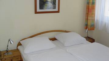 Un lit bien fait, avec du linge de lit blanc, se trouve dans une pièce dotée d’une tête de lit en bois. Un tableau encadré est accroché au mur au-dessus du lit, une lampe de chevet est posée sur la table de chevet, et une fenêtre est ornée d’un rideau.