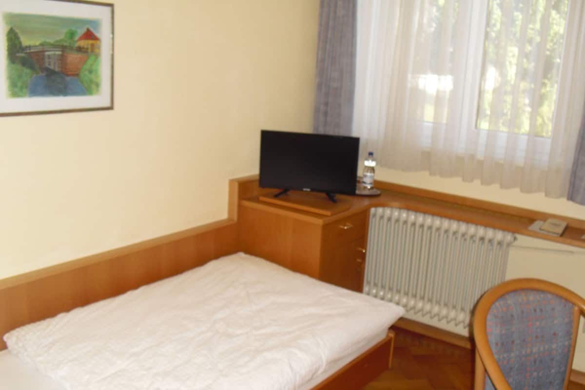 Una habitación de hotel pequeña con una cama individual, un aparador de madera, un televisor encima, una silla y una ventana con cortinas.