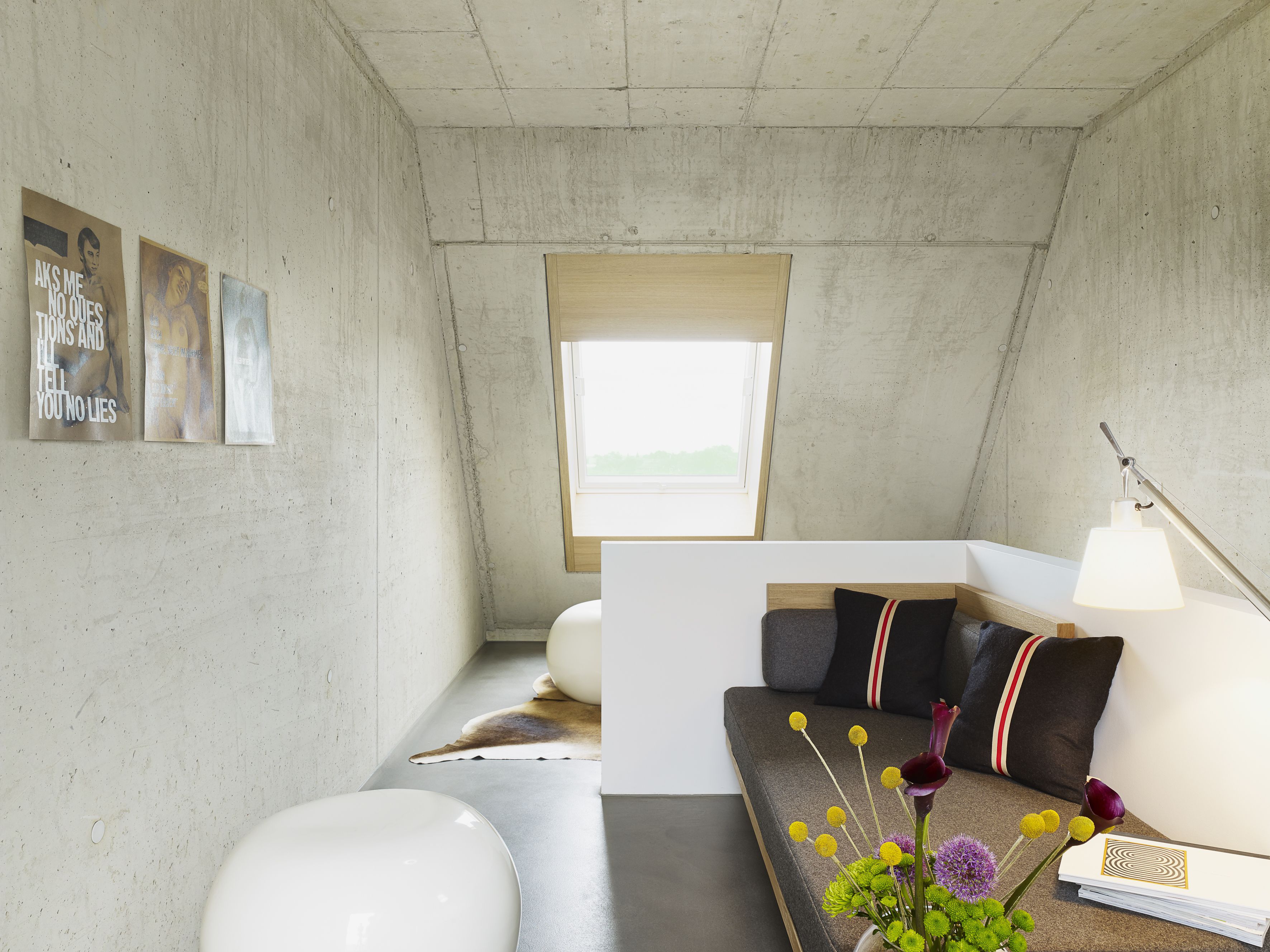 A modern, minimalist room with a concrete wall, a window, a sofa, a lamp, and a vase with flowers.