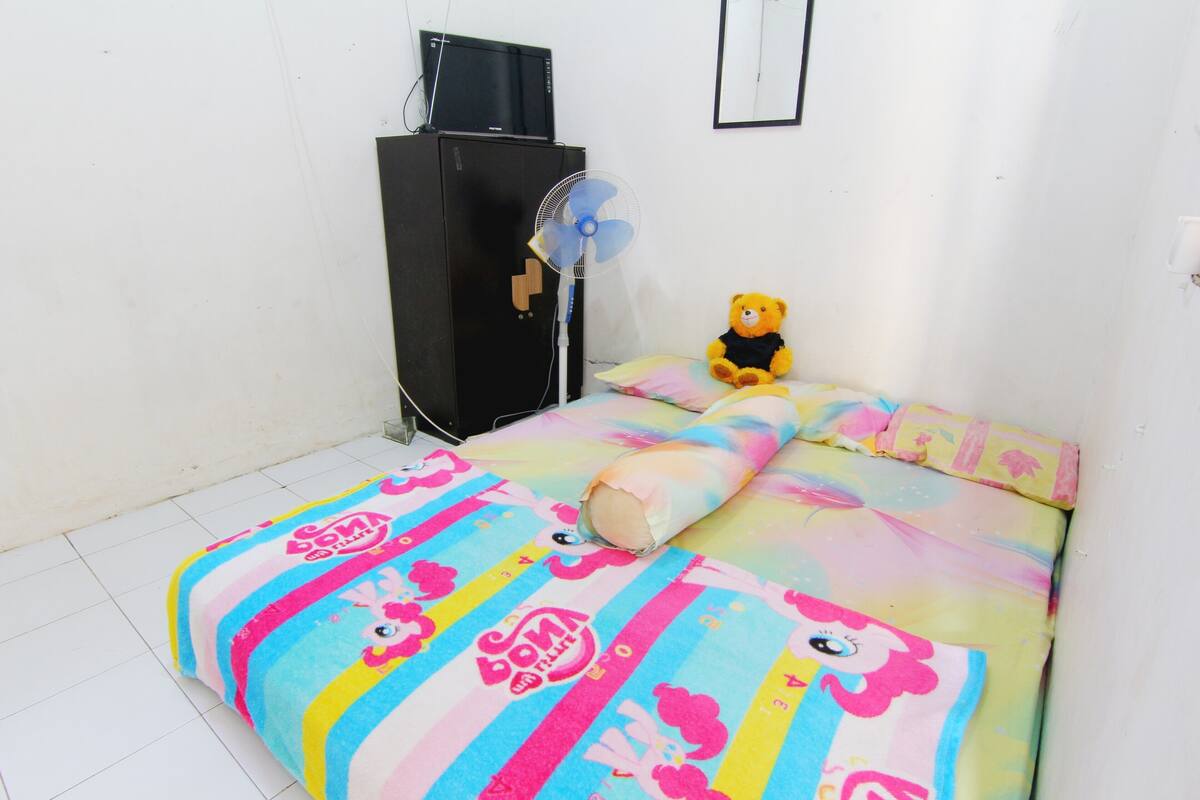 Habitació amb un llit cobert per un llençol colorit, un os de peluix damunt del llit, un armari negre i un ventilador de peu.